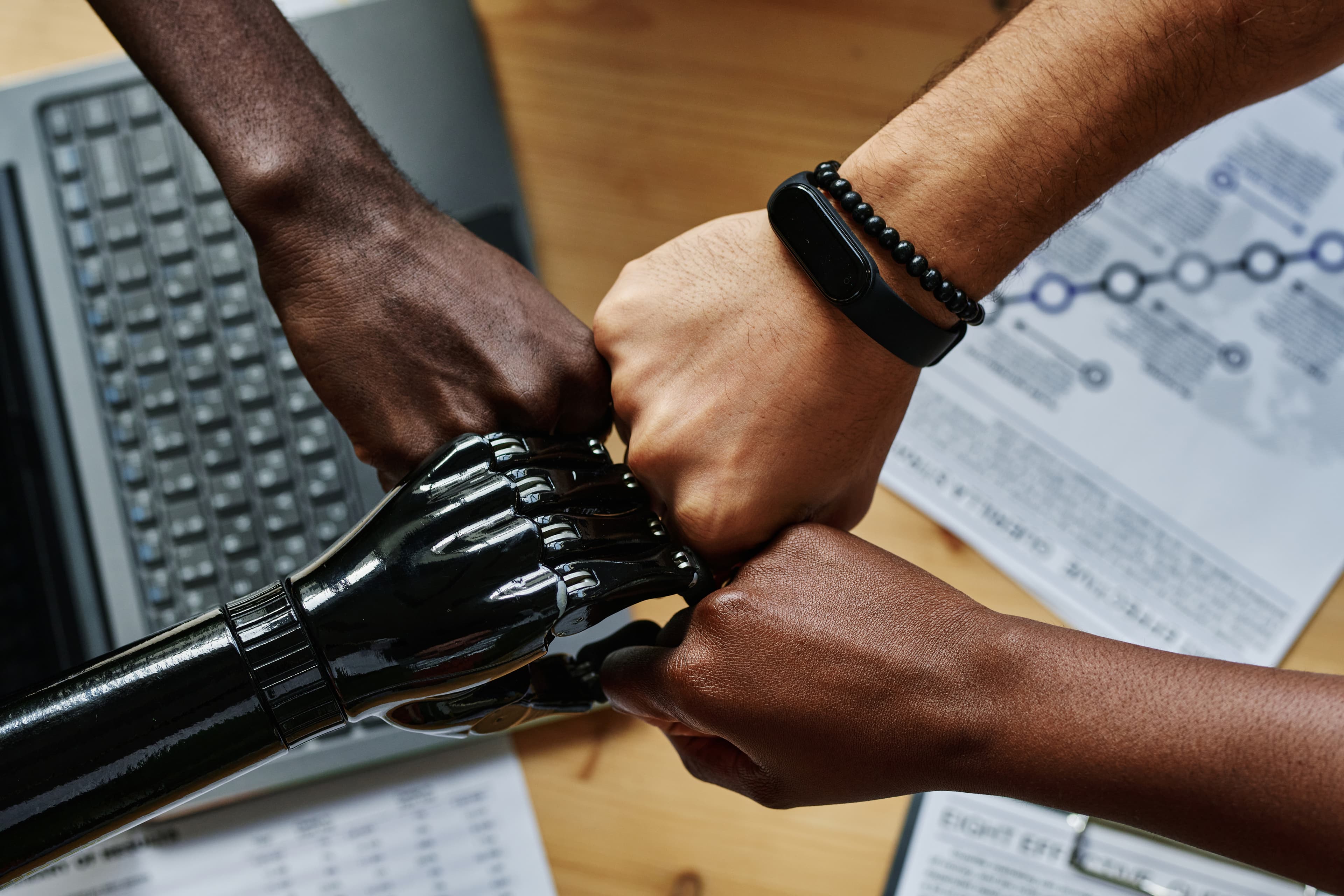 Three human hands and one black robotic hand meeting in unity over a desk — human and AI working as one team